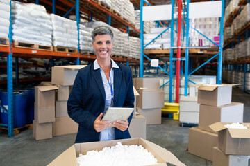 Smiling businesswoman using tablet in warehouse surrounded by boxes and shelves, copy space