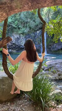 A beautiful girl in dress sits on a thick liana and swings overlooking the beautiful Ponytail Falls along the Tully River and the dense rainforest. Tropical North Queensland, Tully, Australia