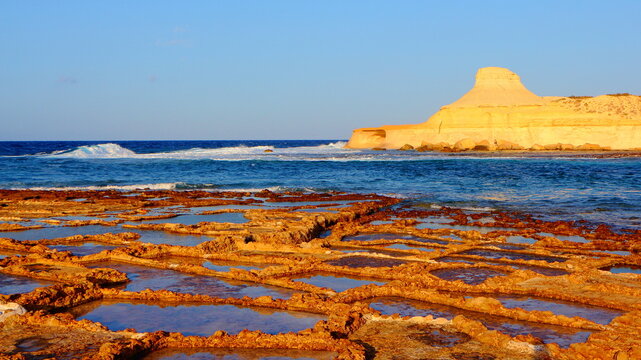 Salt pans, salterns or salt evaporation ponds in the Gozo afternoon sun with blue sea. Orange yellow red limestone rocks and stone shapes. Sea waves and deep blue, beautiful ocean shore.