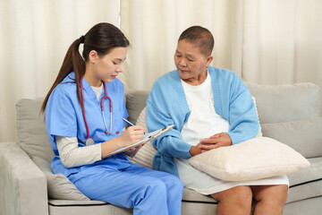 Obraz premium Young adult Caucasian female physiotherapist writing health information on clipboard beside Asian senior female cancer patient wearing white dress and blue sweater sitting on sofa indoors