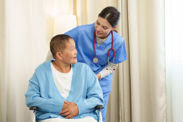 Fototapeta premium Caucasian female physiotherapist in blue medical scrubs standing behind Asian senior female cancer patient sitting in wheelchair smiling during supportive home care therapy session