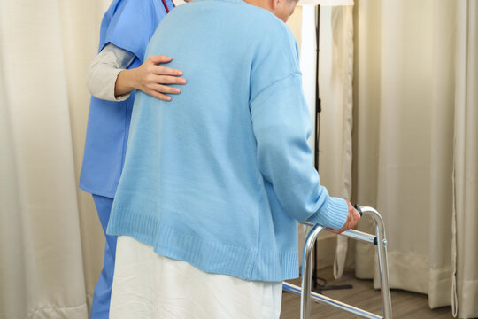 Caucasian young adult female physiotherapist assisting Asian senior female cancer patient in standing up using metal walker inside cozy room during supportive physical rehabilitation therapy session