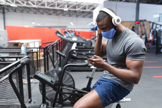 Man wearing mask and headphones using smartphone on exercise bike at gym, copy space