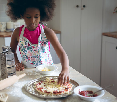 Young girl making homemade pizza in kitchen, adding cheese with focus and care