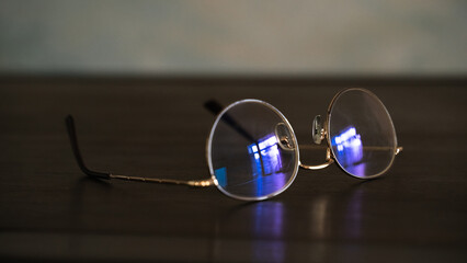 Round eyeglasses with blue light reflection on a dark wooden surface close-up. Vintage style, optical accessory, classic design, minimalism, technology, vision, elegance, retro aesthetic, spectacles.