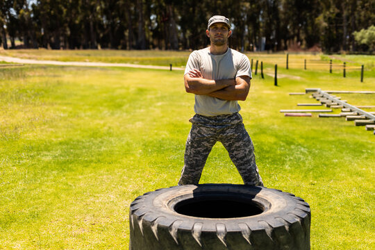Military man in camouflage standing confidently by large tire on training field, copy space