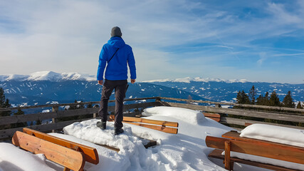 Hiker man with scenic view of snow-capped mountains of Seckauer Alps seen from Steinplan, Murtal, Austria. Snowy benches Wooden terrace of Schutzhaus. Serene alpine landscape in remote Austrian Alps