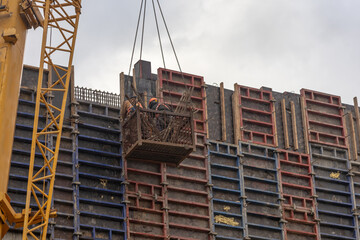 Workers perform wall concreting work while standing in a cradle suspended from crane cables. The technology of concrete wall construction. Reinforcement of walls during construction.