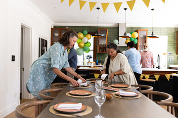 Diverse senior friends preparing table for festive gathering in decorated home kitchen