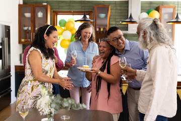 Senior diverse friends celebrating birthday at home, laughing and enjoying wine together