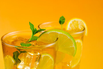 A close-up of two glasses of a ice tea with lime and lemon slices in them. The drink is served in a glass with mint leaves on the top. Warm orange background.