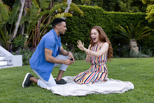 Diverse couple, Young man proposing to excited woman on picnic blanket in lush garden - Powered by Adobe