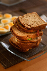 A photo of stacked breakfast sandwiches made with cucumber, cheese, and salami on whole wheat bread, with defocused boiled eggs in the background