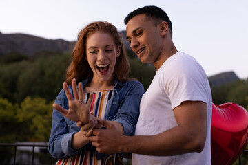 Young diverse couple celebrating engagement in garden, woman admiring ring with excitement