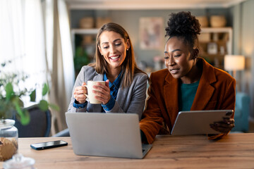Two Diverse Women Collaborating on Startup Ideas in a Bright Home Office Setting with Laptops and Tablets