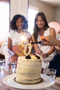 Lighting candles on birthday cake while diverse female friends celebrate at home, smiling joyfully