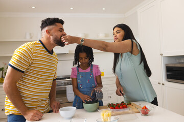 Mother feeding husband while daughter prepares fruit salad in kitchen together
