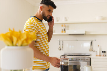 In kitchen, man talking on smartphone while making coffee, looking focused