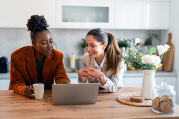 Two Professional Women Laughing While Working Together On Laptop In Bright Modern Kitchen Setting
