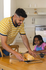 Father slicing bread at kitchen table while daughter eagerly watches, smiling