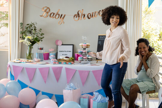 diverse female friends celebrating baby shower with gifts and colorful decorations, at home