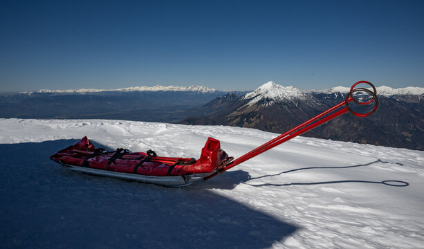 Red rescue sled on a snow-covered mountain peak under bright sunlight with a backdrop of majestic alpine ranges and a clear blue sky