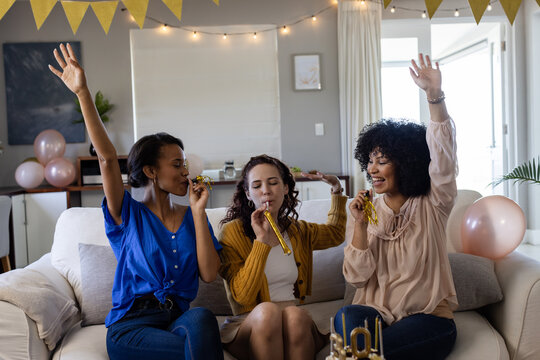 Celebrating at home, diverse female friends with party blowers and decorations, having fun together