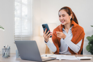 Asian businesswoman smiling while using smartphone and working on laptop in bright office setting.