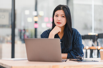 Focused Asian businesswoman working on laptop in modern office, expressing determination and...