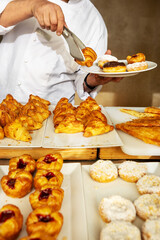 Close-up of chef in white uniform serving fresh pastries with tongs at bakery buffet or in restaurant of hotel. Assortment of croissants, donuts and sweet pastries on display