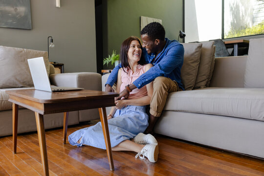 Diverse couple sitting on floor at home, smiling and enjoying time together