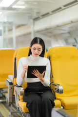 Asian Woman on Plane: An elegant Asian woman is engrossed in using a tablet, seated in a brightly...