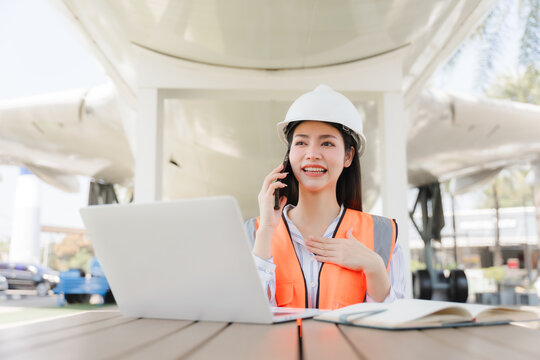 Aviation Engineer: A skilled female aviation engineer, donned in a safety helmet and vest, manages operations on her laptop while speaking on the phone with a cheerful expression.
