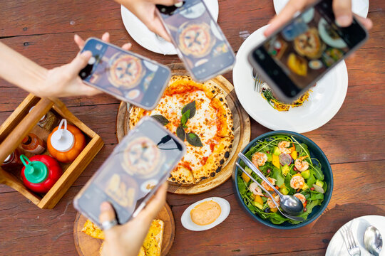 Group of Happy Asian millennial people friends enjoy and fun celebration reunion meeting outdoor dinner party using mobile phone taking picture of food and drink on dining table at restaurant garden.