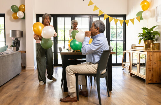 Diverse senior friends preparing for celebration, inflating colorful balloons, at home - Powered by Adobe
