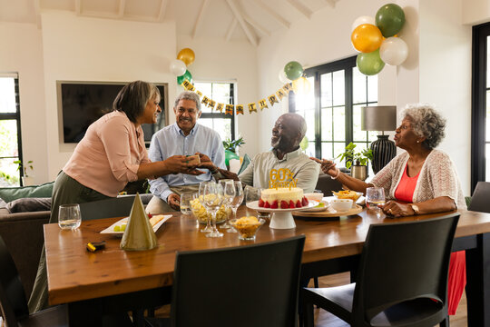 Diverse senior friends celebrating 60th birthday with cake and decorations at home, sharing joy - Powered by Adobe