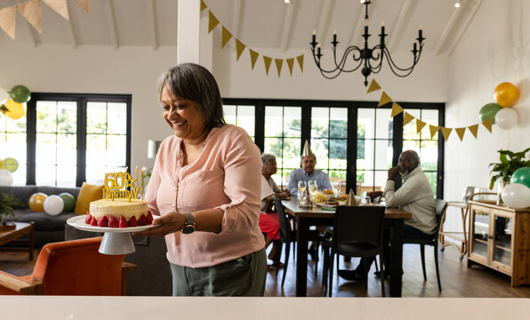 Woman carrying birthday cake with diverse senior friends celebrating in bright living room, at home