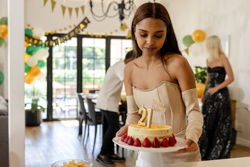 Young woman carrying birthday cake with 21 candles at party, at home