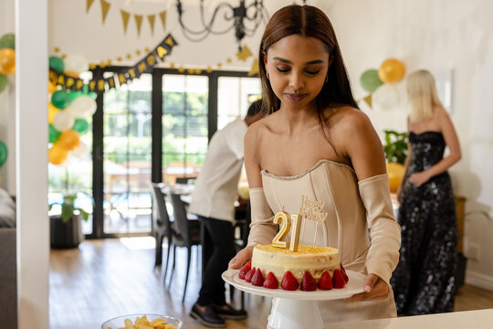 Young woman carrying birthday cake with 21 candles at party, at home