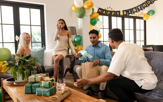 Birthday, diverse friends opening gifts and having drinks in party living room, at home