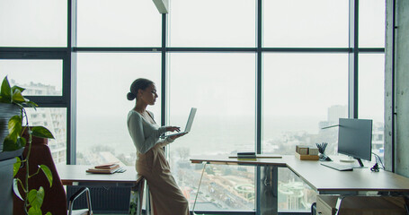 A young professional woman working on a laptop in a modern office with large windows and a scenic...