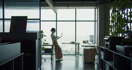 A young professional woman working on a laptop in a modern office with large windows and a scenic view, embodying focus, productivity, and a contemporary workspace atmosphere