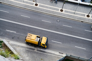 A yellow garbage truck is picking up waste along an urban street with pedestrians strolling in the...