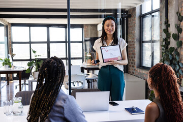 Asian woman presenting award to diverse female colleagues in modern office, smiling confidently