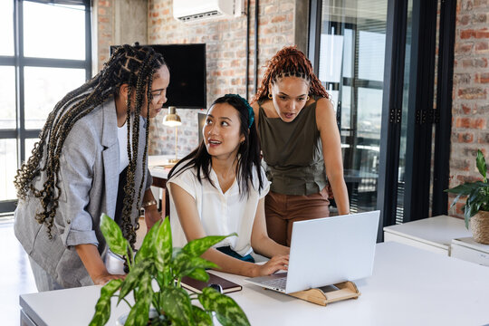Collaborating in office, diverse female team sharing ideas around laptop enthusiastically