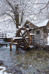 Fototapeta premium A snow-covered watermill stands beside a flowing stream, its wooden wheel contrasting with the stone hut and winter landscape, capturing a rustic, serene scene.