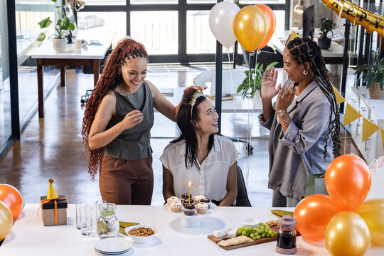 Female diverse colleagues celebrating birthday with cupcakes and balloons in modern office