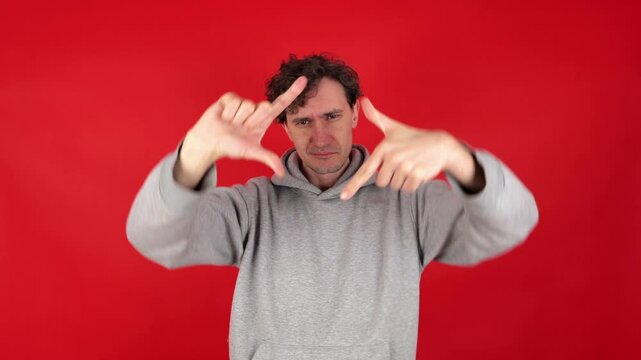 young adult curly man in casual grey hoodie making photo frame gesture with hands, filled creativity imagination. standout photography concept. Isolated red background studio shot. creativity in focus