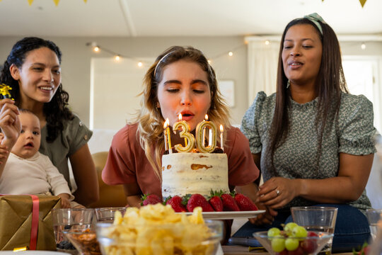 Woman blowing out candles on birthday cake surrounded by diverse female friends at home