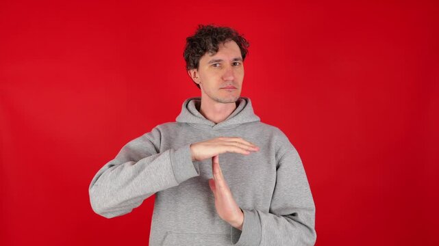 young adult curly serious man holds hands in front of shows sign timeout gesture. wearing grey hoodie. isolated red background studio shot. Concept - pause, break, rest, vacation